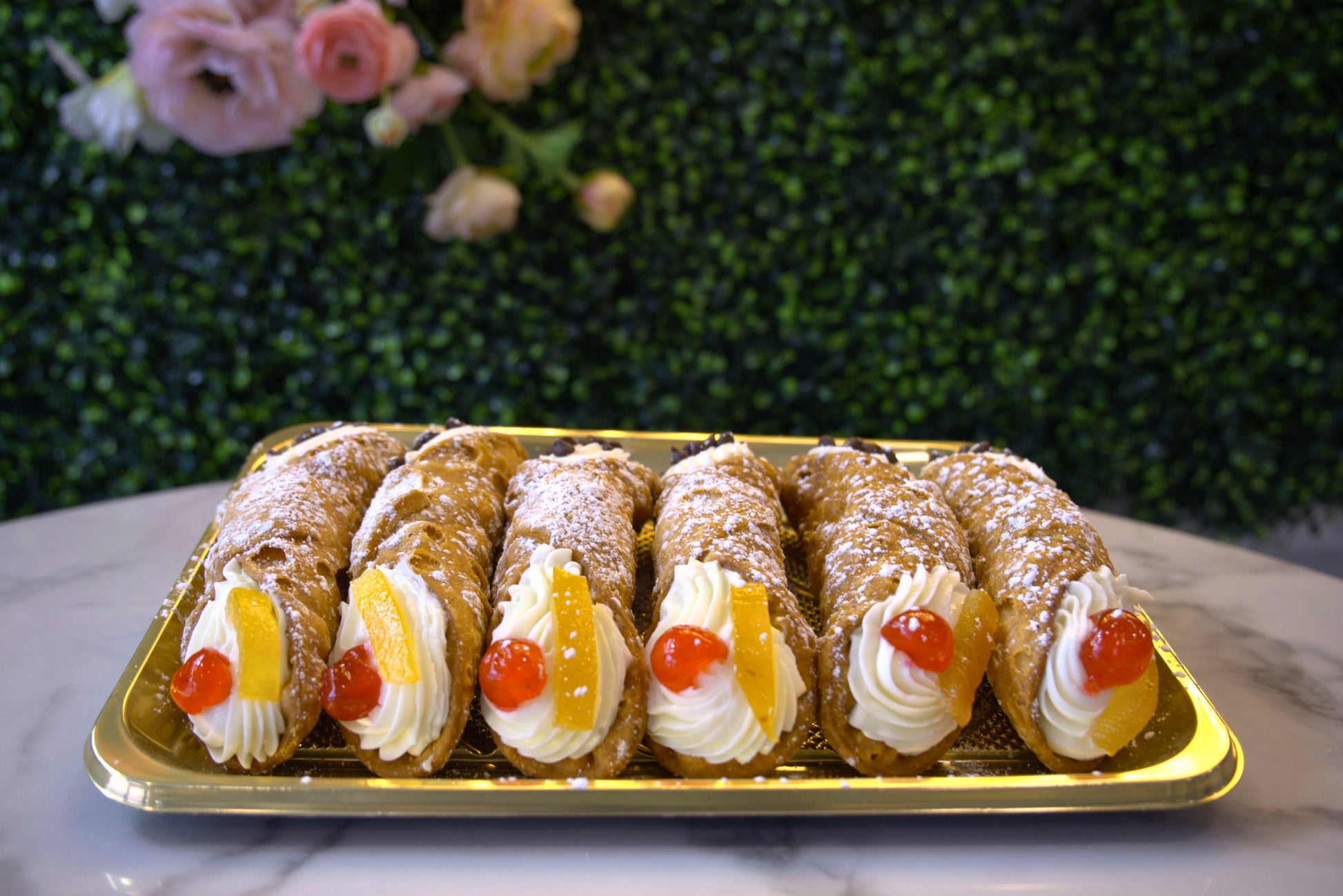 The fruit side of a tray of cannoli in front of greenery and flowers from La Roma Pastry Shop