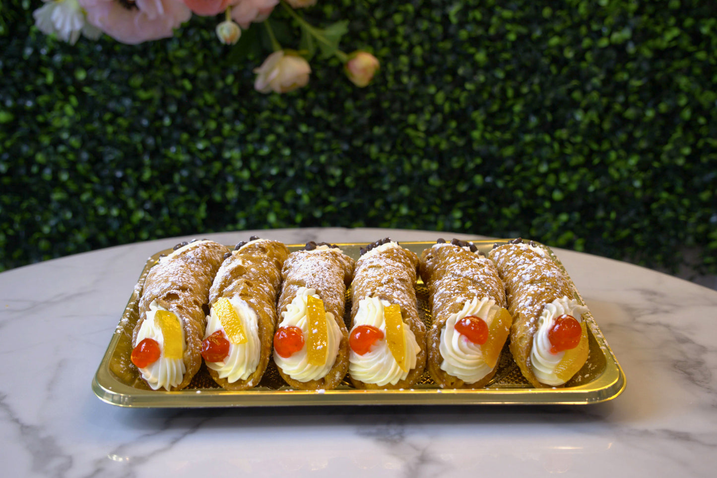 Display of a half dozen cannoli in front of floral greenery made by La Roma Pastry Shop