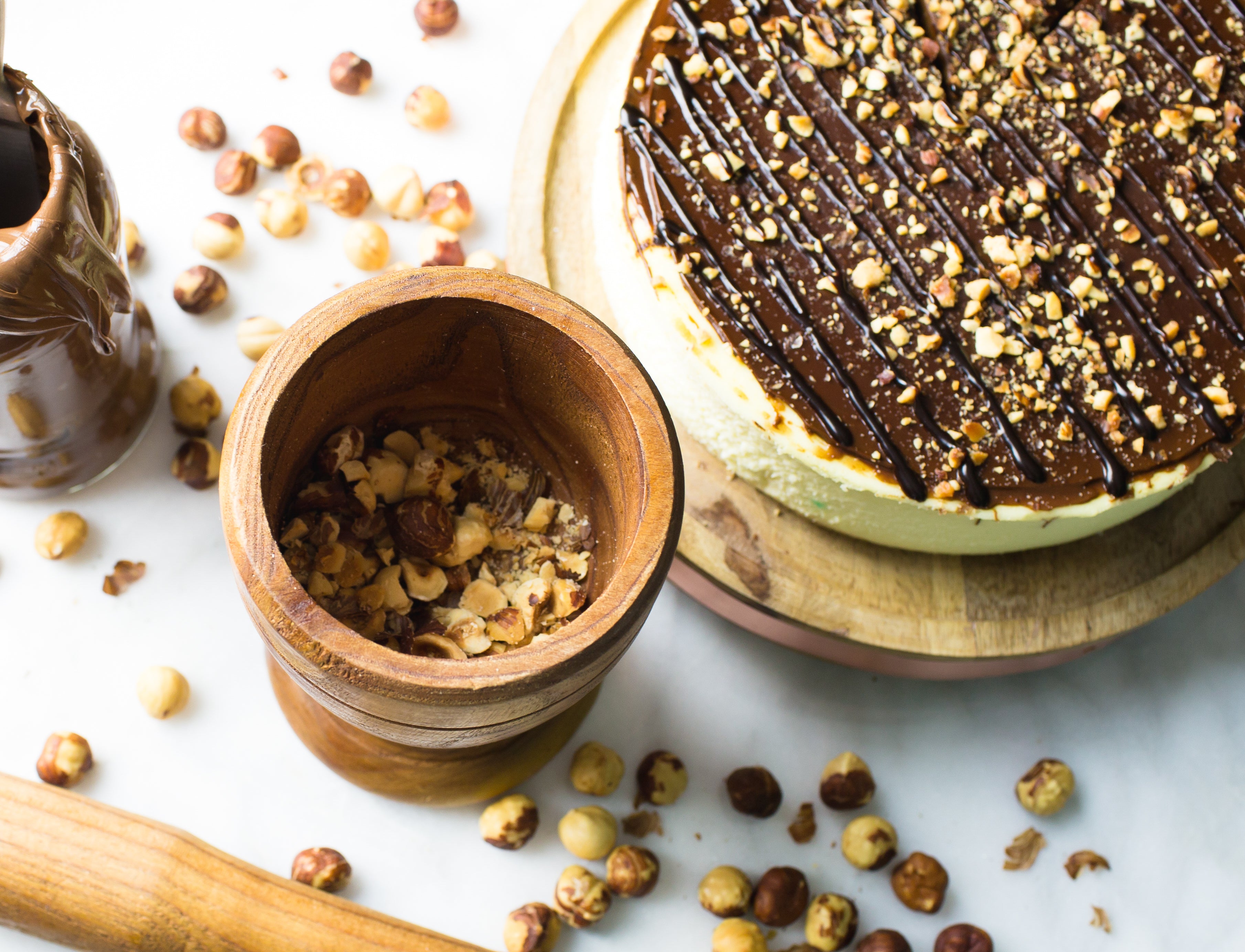 overhead image of nutella cheesecake with hazelnuts and chocolate on top by la roma pastry shop in oakdale new york with more hazelnuts scattered around and pieces in wooden mortar with pestle laying nearby 