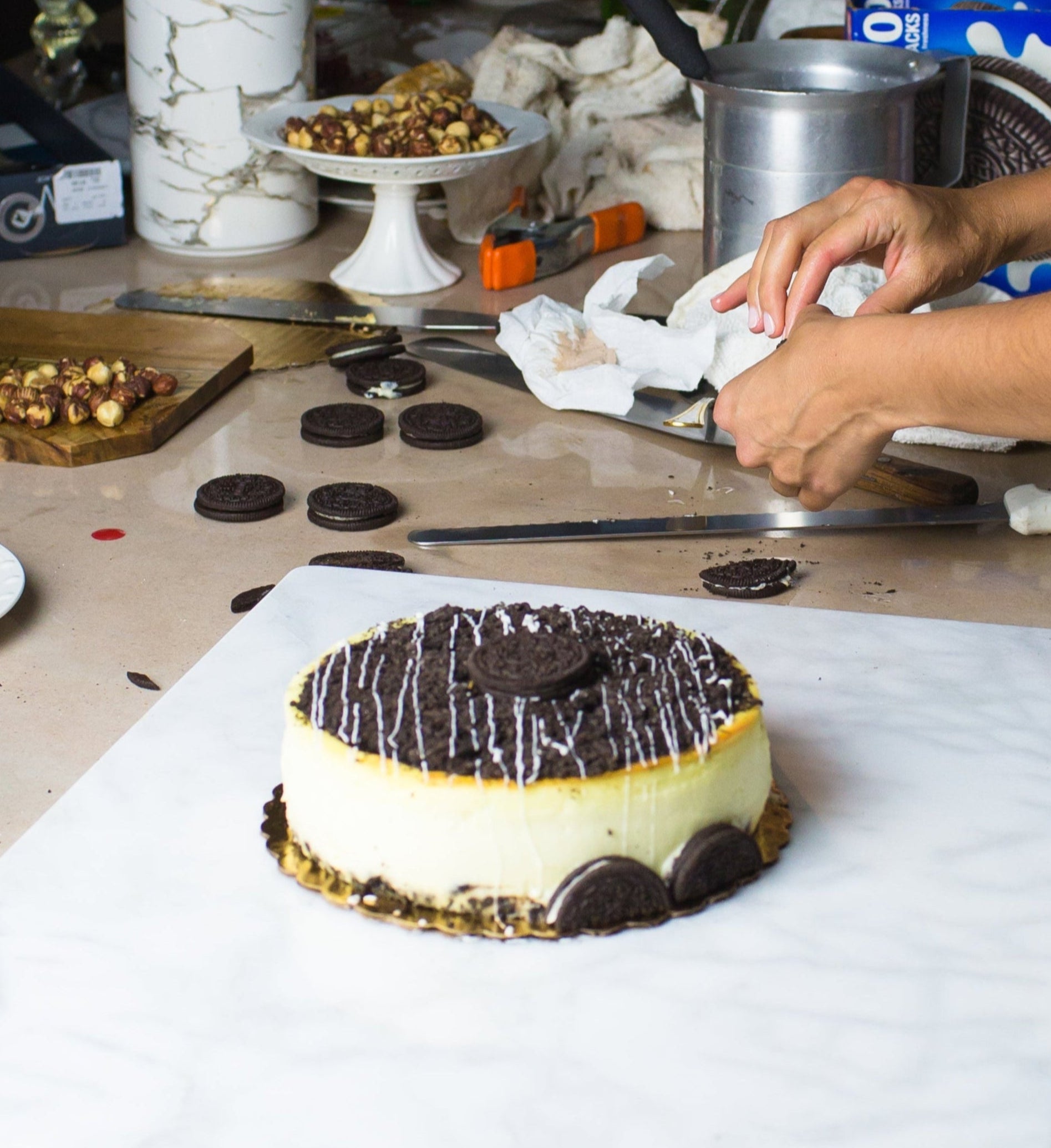 oreo cheesecake made by la roma pastry shop in new york sitting on marble board with hands preparing oreos nearby and hazelnuts and other props scattered around