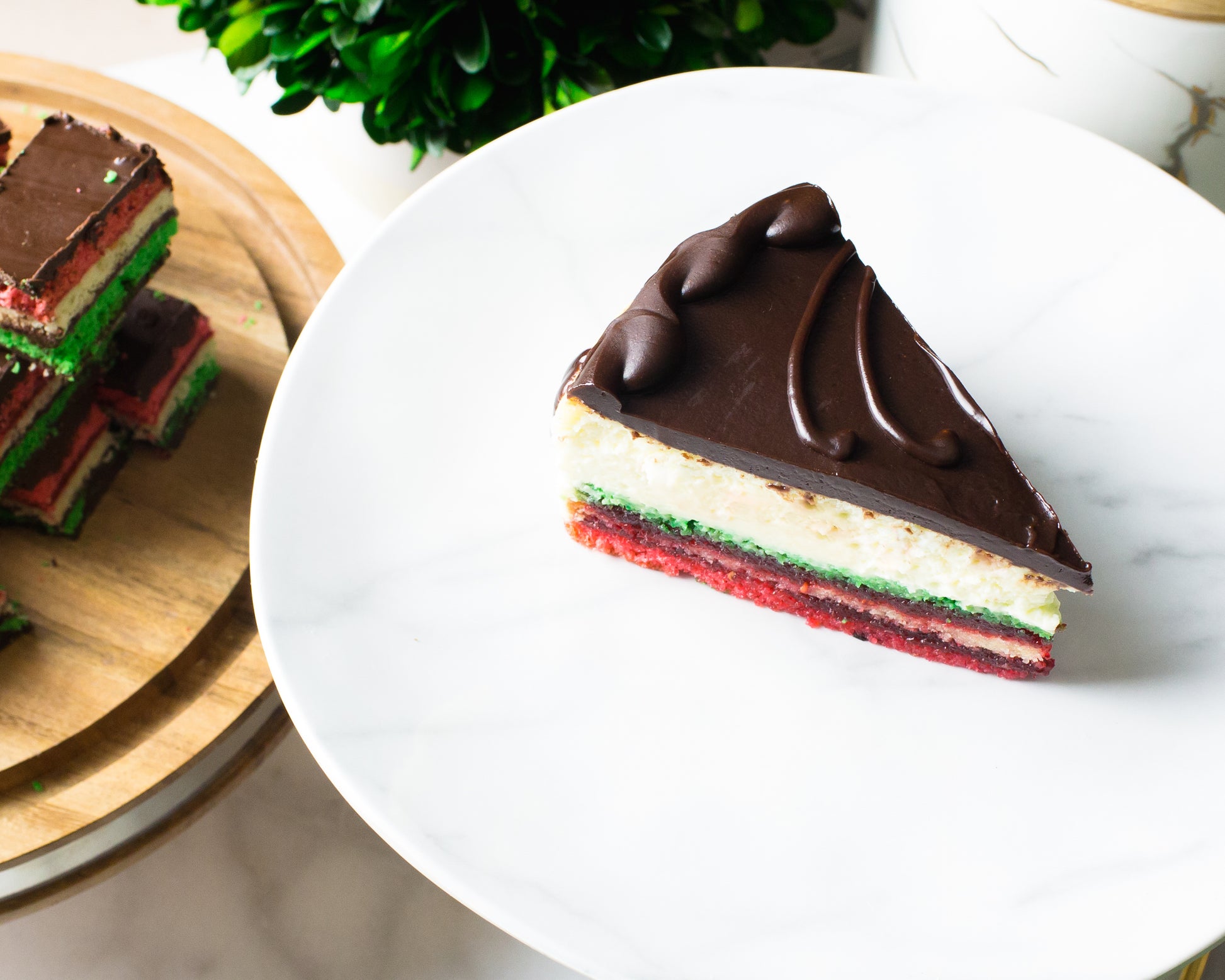 slice of rainbow cookie cheesecake on marble cake stand with chocolate frosting baked by la roma pastry shop in new york